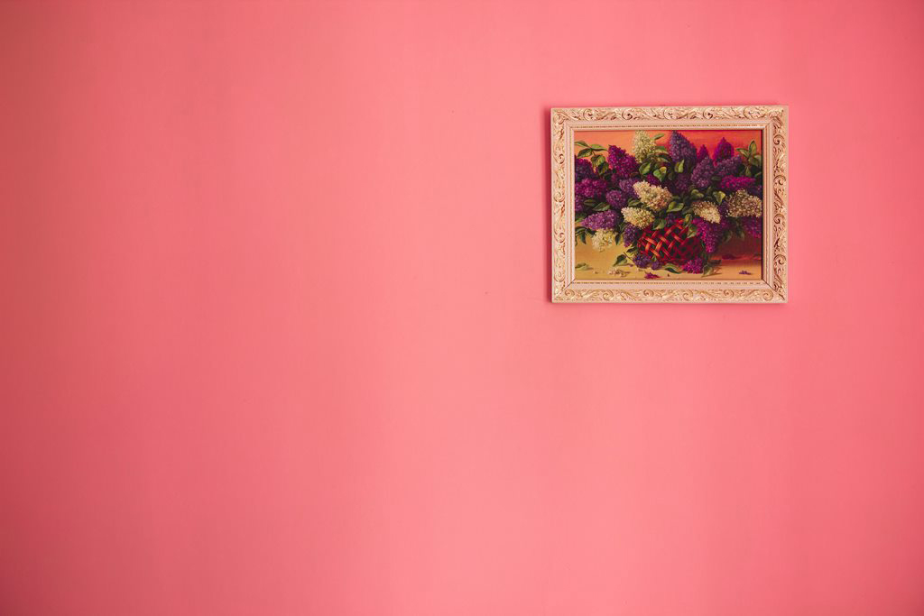 An interior wall painted a vibrant beautiful pink with a hanging framed floral painting on the right side.