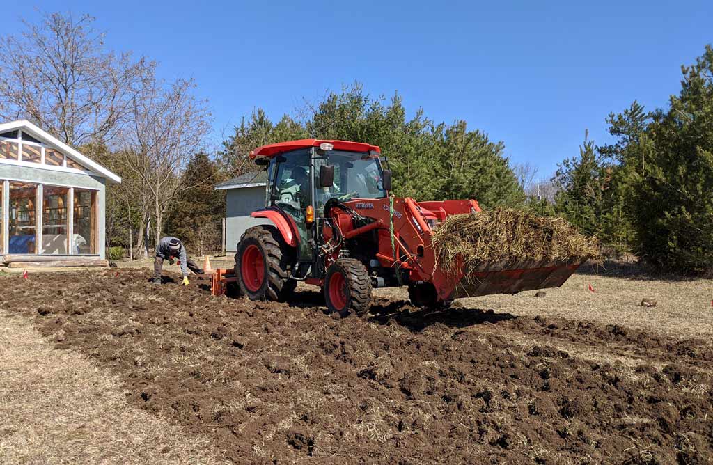 A large red tractor tilling a new garden bed and Justin collecting rocks from the soil.