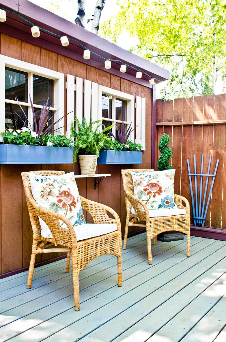 A backyard patio with two rattan chairs, flanked by an outdoor shed with bright blue window boxes.