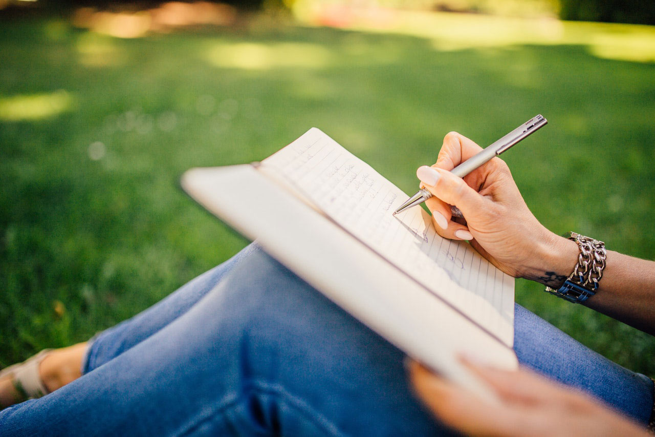 A woman sitting on the grass, writing in a journal which is on her knees.