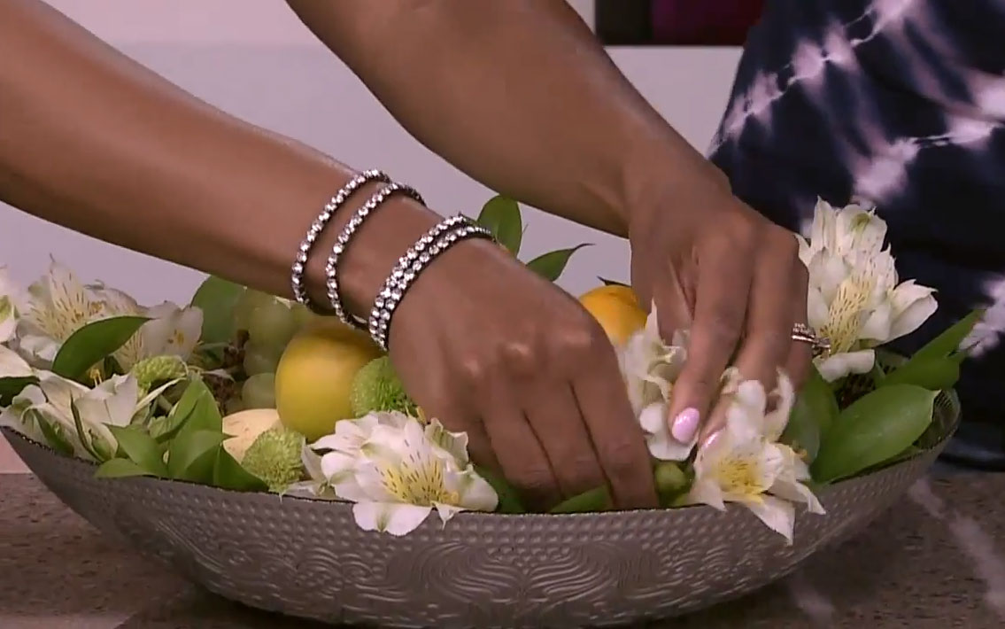 A close up of Tracy Moore’s hands adding florals to a bowl of fruit and greenery.