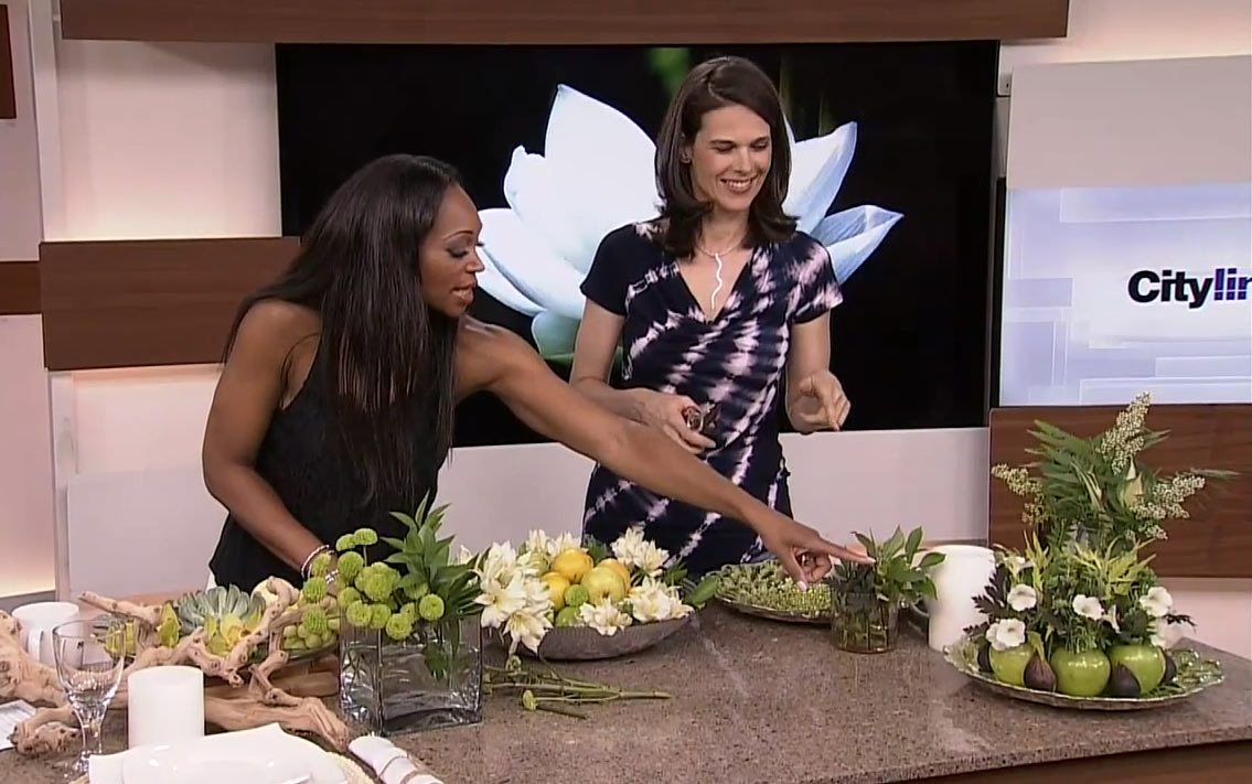 Celia Alida Rutte and Tracy Moore are pointing to a beautiful fruit and floral arrangement, on the set of Cityline.