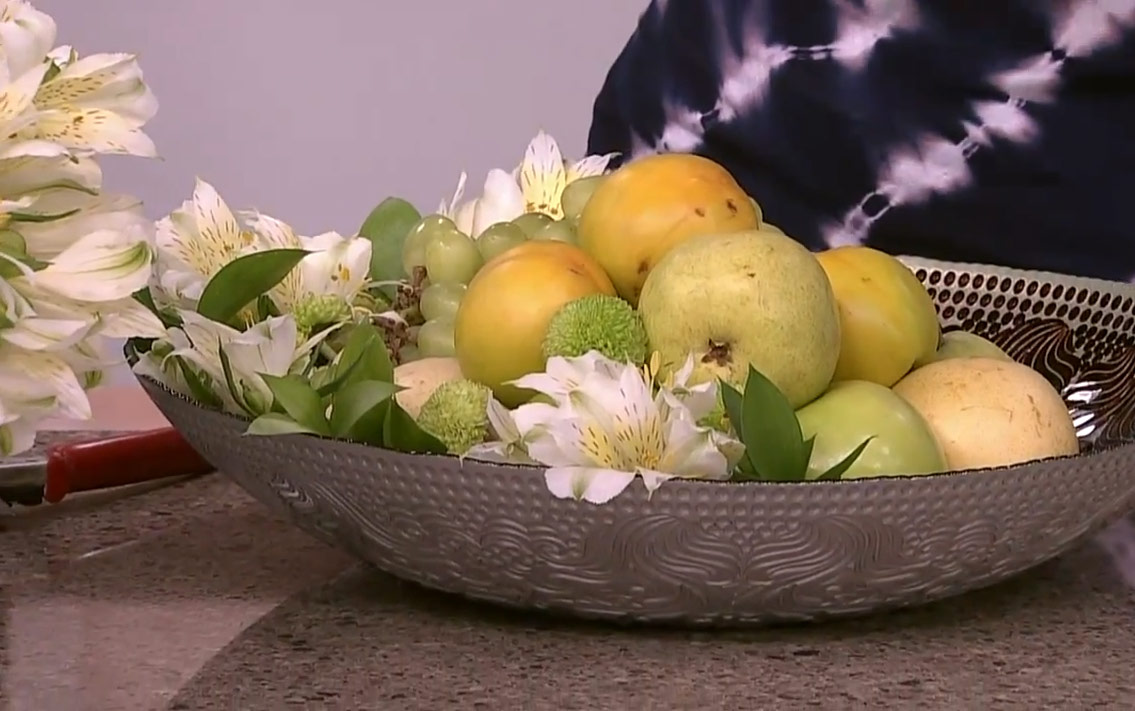 A bowl of fruit, flowers and greenery arranged in a wide bowl.