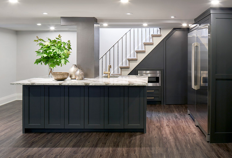 A basement kitchenette with white counter and dark grey cabinets built into the stairs and surrounding area making maximum use of the space.
