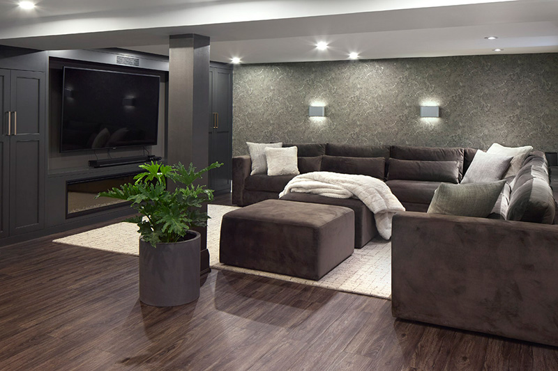 A large dark brown sectional with off white pillows, an off white area rug, and dark brown floors, in a theatre room with dark grey cabinets and a large TV.