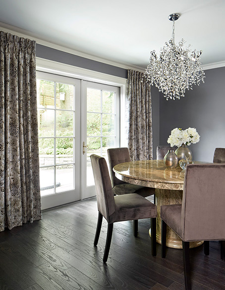 A dining room designed in shades of purple grey, with a large round stone table, dining chairs, floor to ceiling drapery and a chrystal chandelier.