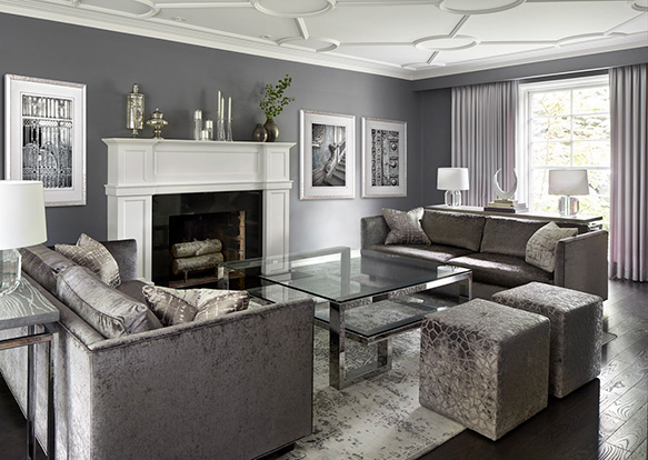 A living room designed in shades of purple grey, with two sofas, two square ottomans, a large square glass coffee table, and a large white fireplace mantle.