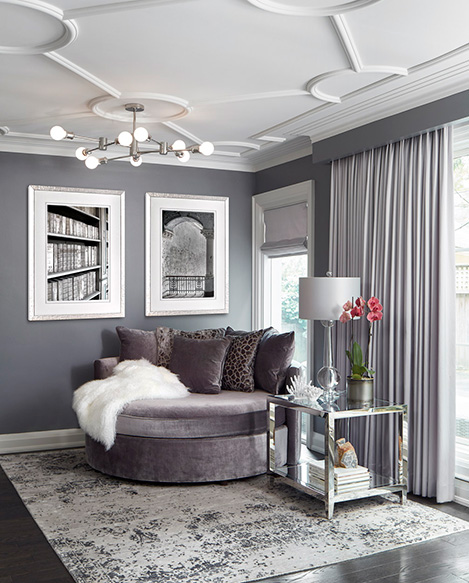 A reading nook designed in shades of purple grey with artwork on the walls, a large round reading chair, a side table, floor to ceiling drapery and an area rug.